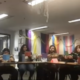 A group of people sitting around a table, each holding up a colorful sheet of paper, in a room decorated with rainbow flags.